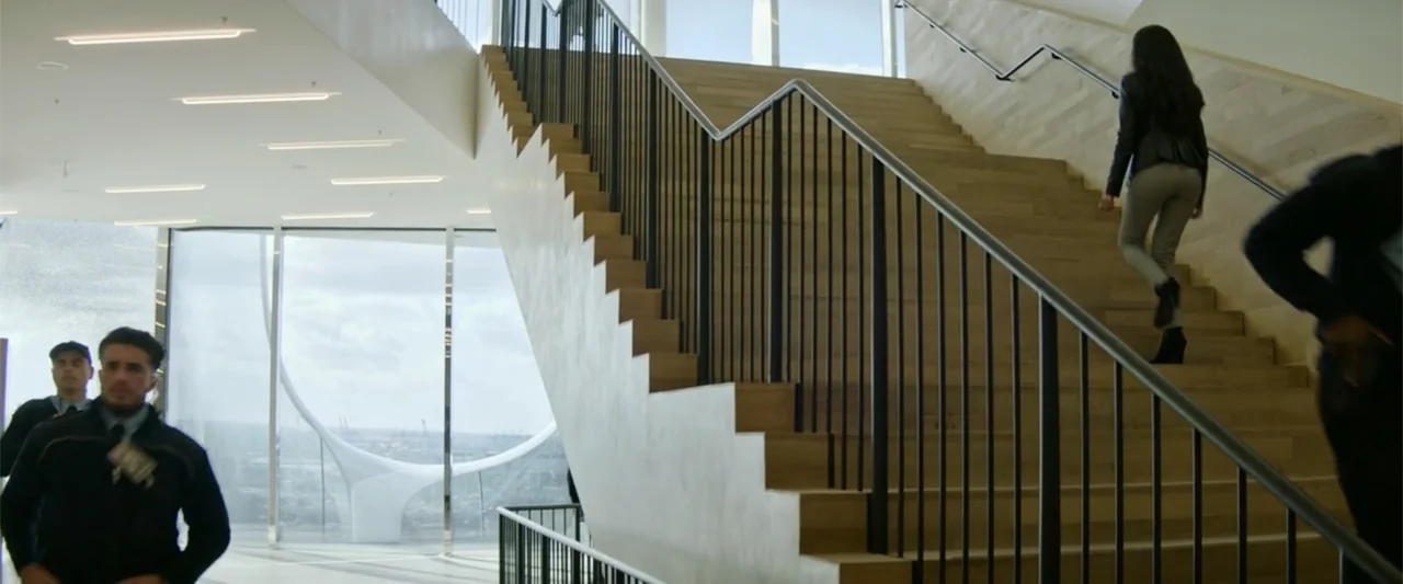 Charlie's Angels (2019) movie still: wide — A modern glass-walled lobby features a central wooden staircase with black metal railings. A woman…
