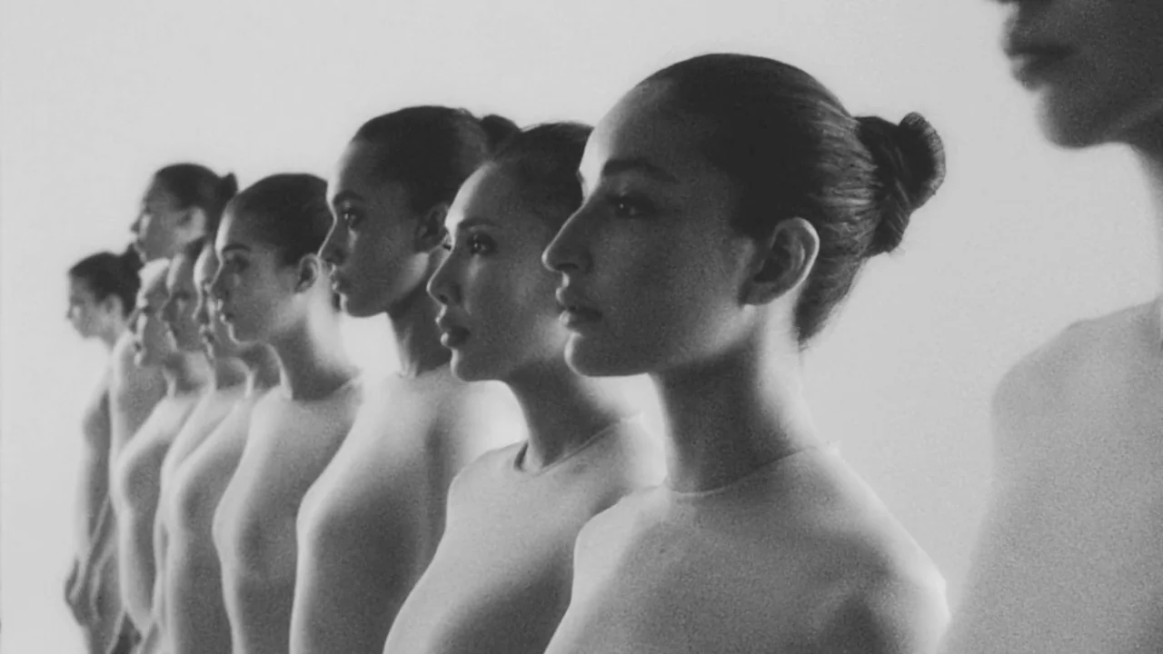 Divinity (2023) movie still: medium — A row of identical women stands in profile against a plain white background. They all have their hair pulled…