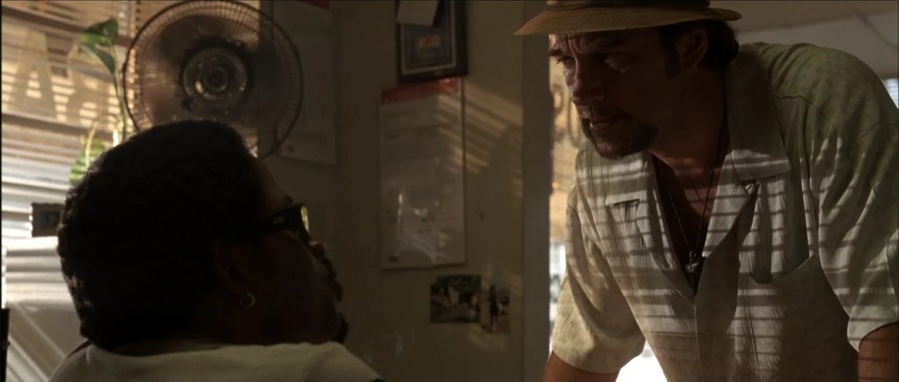 Gang Related (1997) movie still: medium — A cluttered small office features a wall with posted papers and a metal fan. A man in a hat leans over the…