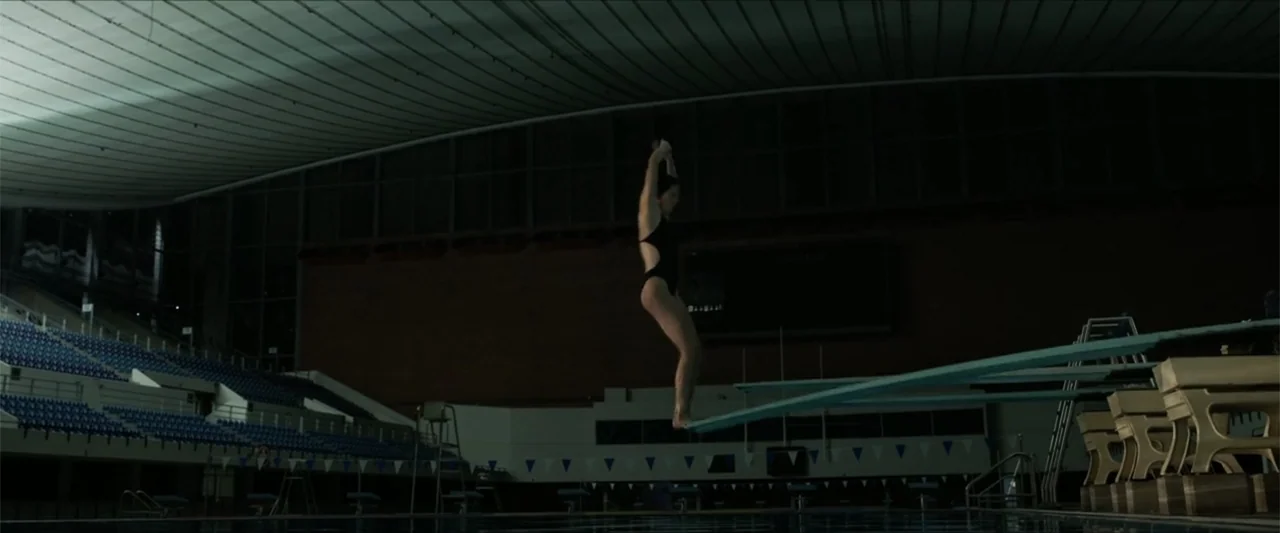 Dive (2013) movie still: wide — A dark, empty indoor aquatic center features tiered seating and multiple long diving boards. A lone female diver in…