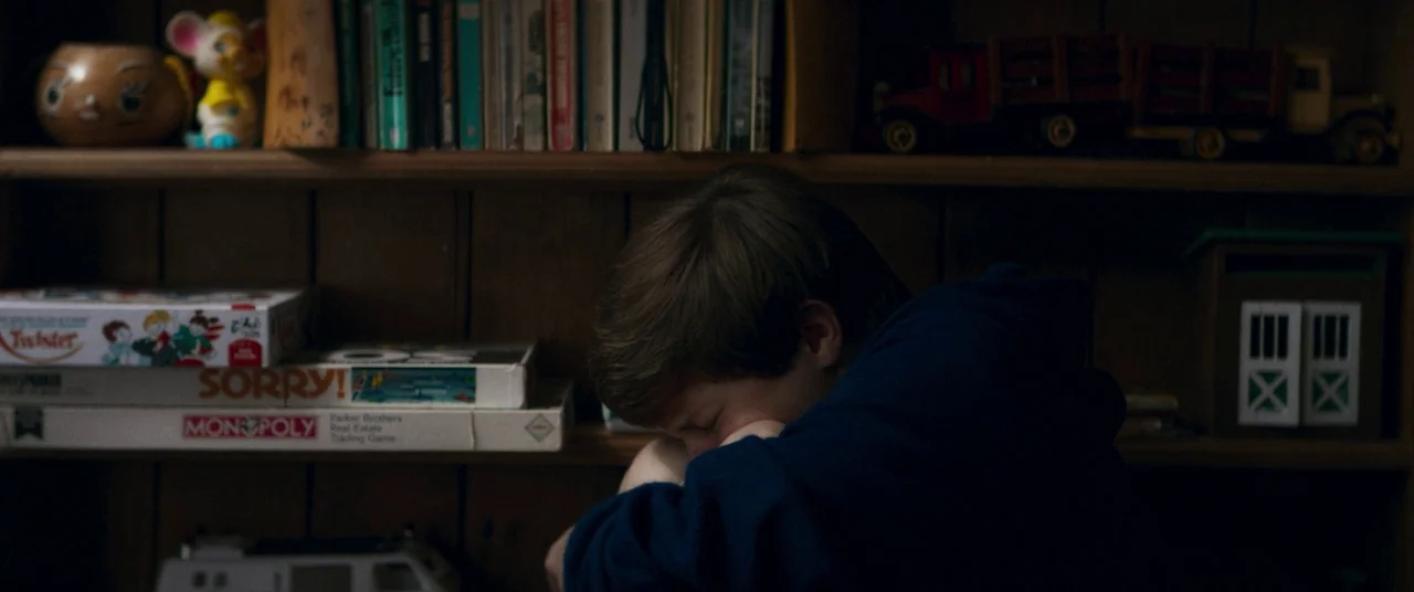 Summer of 84 (2018) movie still: medium — A wooden shelf filled with vintage board games, books, and toy figures. A young person in a hoodie sits…