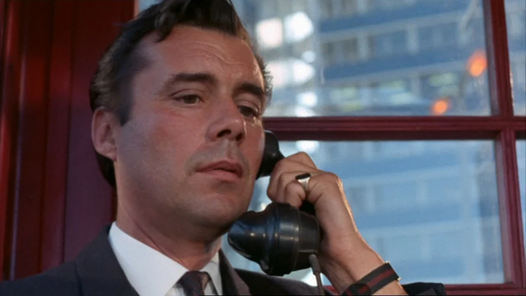 Accident (1967) movie still: closeup — A man stands inside a red-framed telephone booth with a view of city buildings. He speaks into a vintage…
