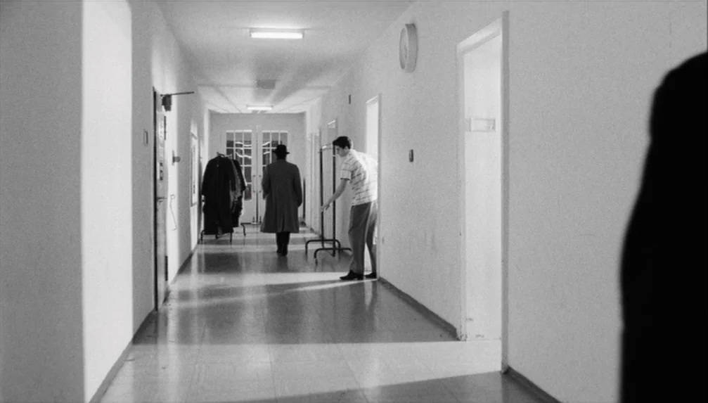 Veronika Voss (1982) movie still: wide — A long, sterile hospital corridor features white walls, linoleum floors, and multiple closed doors. One man…