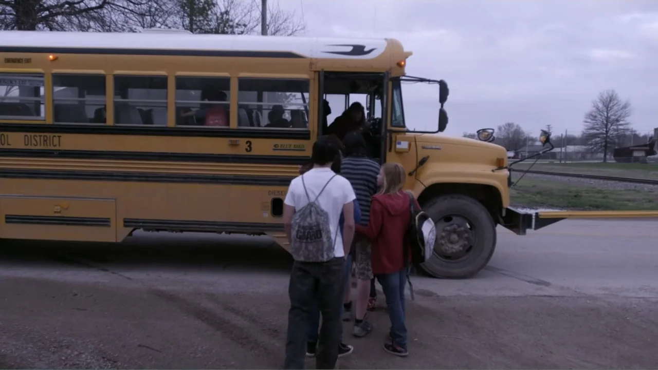 Rich Hill (2014) movie still: wide — A group of teenagers stands near the open door of a yellow school bus. The students are about to climb the…