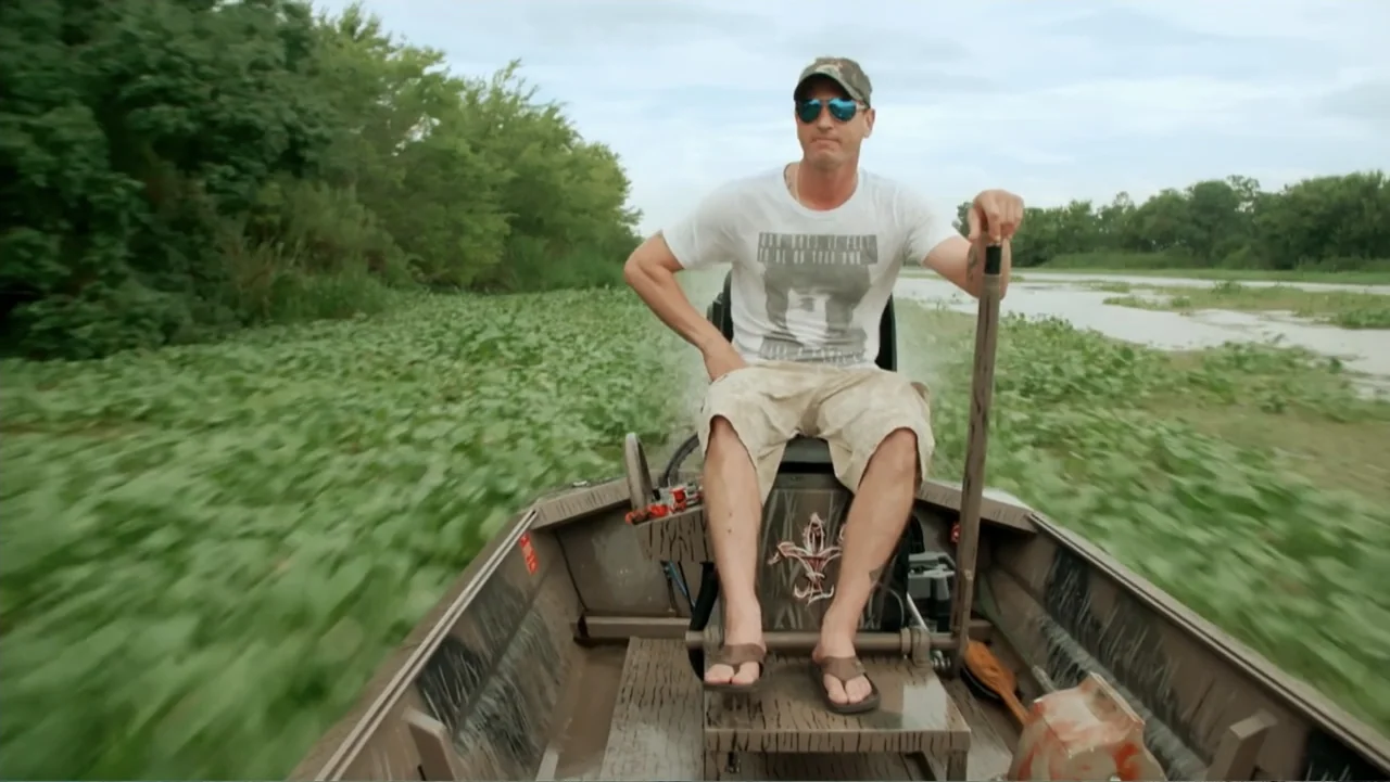 Mommy Dead and Dearest (2017) movie still: medium — A man navigates a camouflaged boat through a marshy waterway covered in lily pads. He sits in…