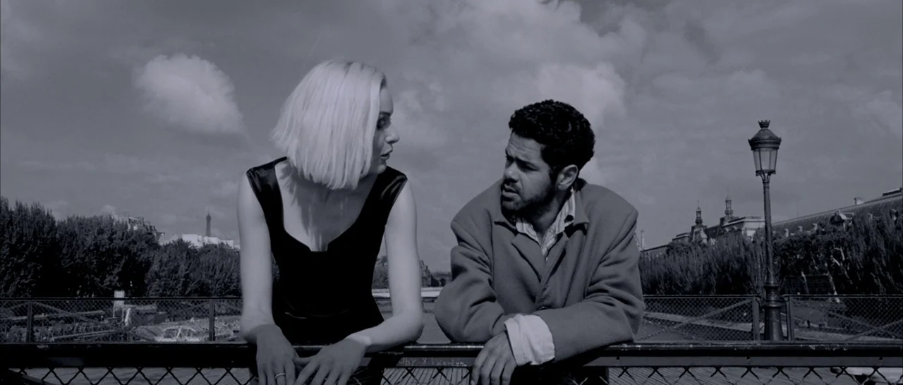 Angel-A (2005) movie still: medium — An iron railed pedestrian bridge over a river with cloudy skies above. A woman and a man face each other while…