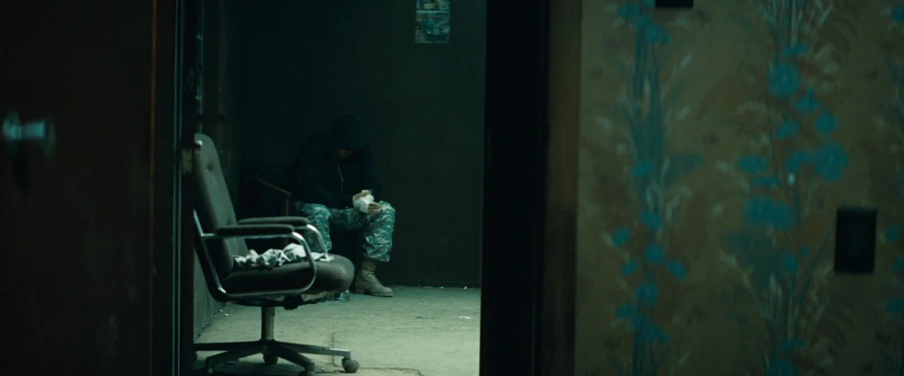 Out of the Furnace (2013) movie still: medium — A dark, sparsely furnished room is viewed through a partially open doorway. A man sits on a chair in…