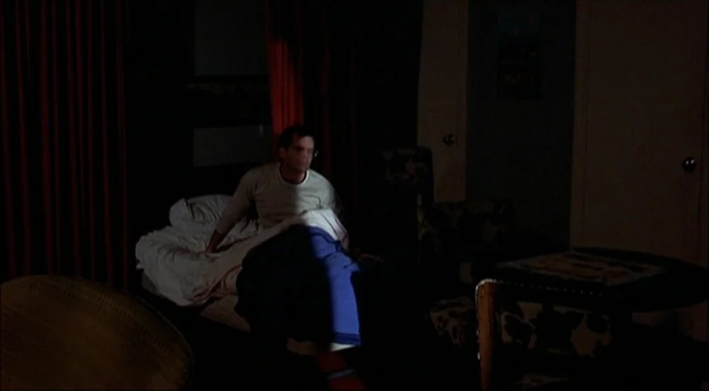 The King Of Marvin Gardens movie still: wide — A dimly lit 1970s motel room features heavy dark curtains and a simple round table. The subject sits…