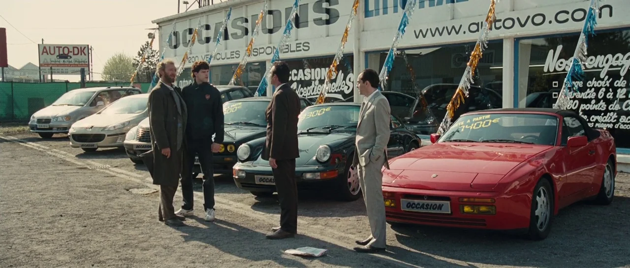 Our Day Will Come (2010) movie still: wide — A gravel lot filled with various cars sits in front of a white dealership building. Four men stand…