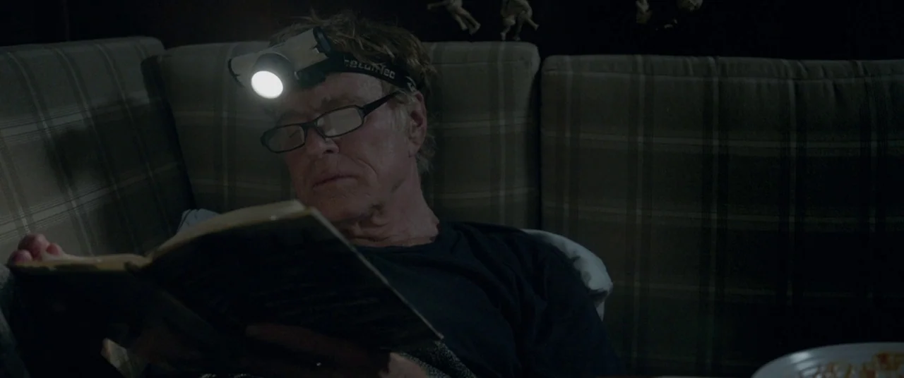 All Is Lost (2013) movie still: medium — An older man sits on a plaid sofa in a dark room. He reads a book illuminated by the bright beam of a…