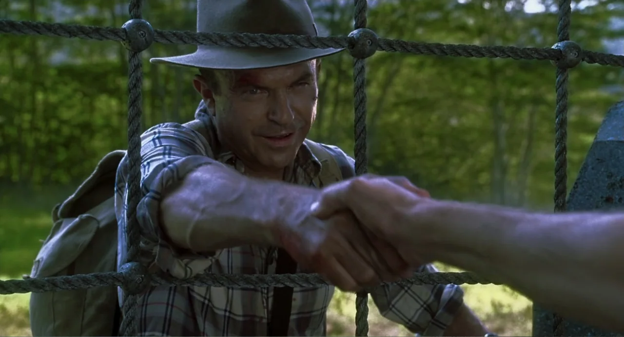 Jurassic Park III (2001) movie still: medium — A man in a fedora reaches through a rope fence in a green park. He is shaking hands with an unseen…