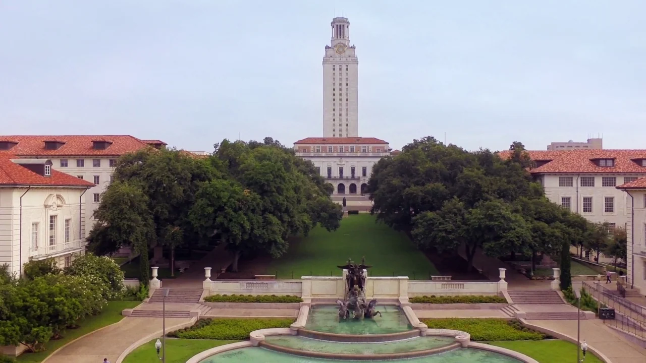 Tower (2016) movie still: establishing — A wide view of a campus mall featuring a large central fountain and tower. Lush green trees line the…