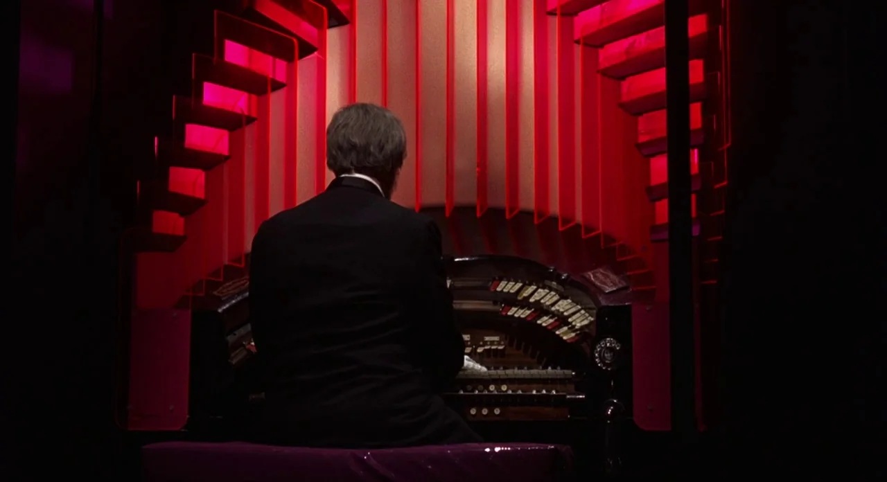 The Abominible Dr. Phibes (1972) movie still: medium — A man sits at a complex pipe organ console surrounded by illuminated red baffles. The man…
