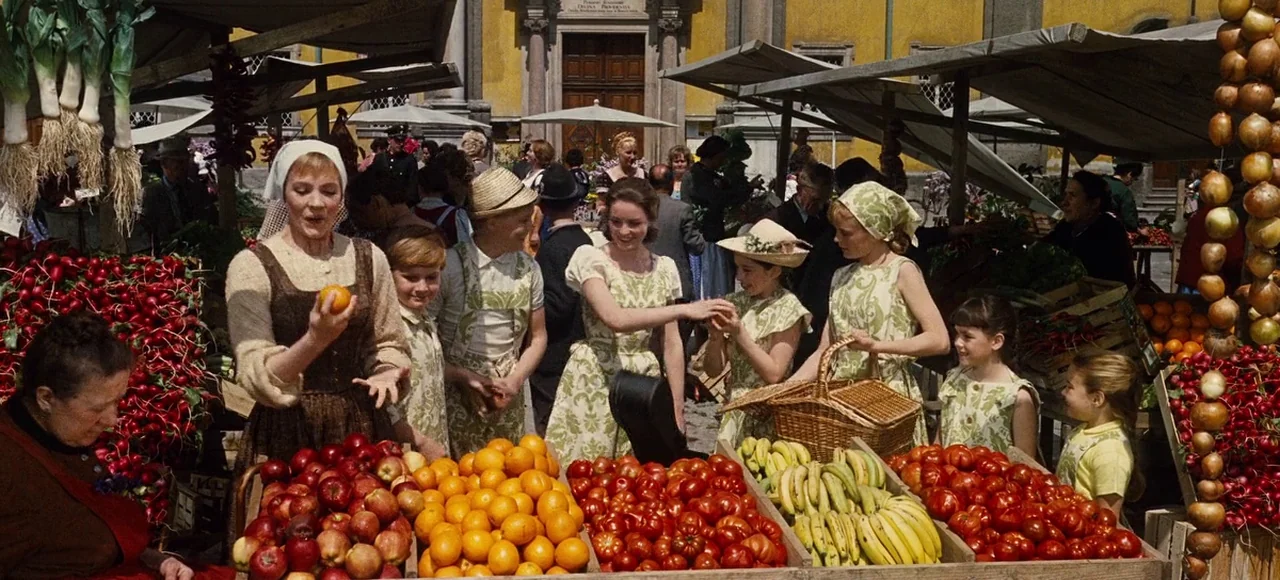 The Sound of Music (1965) movie still: wide — A bustling outdoor produce market features wooden stalls filled with fresh fruits and vegetables. A…