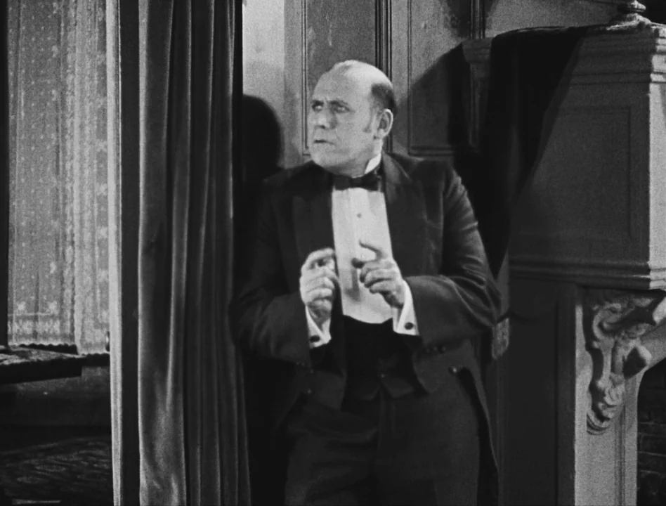 Sherlock Jr. (1924) movie still: medium — A 1930s interior features wooden wainscoting and heavy curtains near a stone fireplace. A man in a tuxedo…