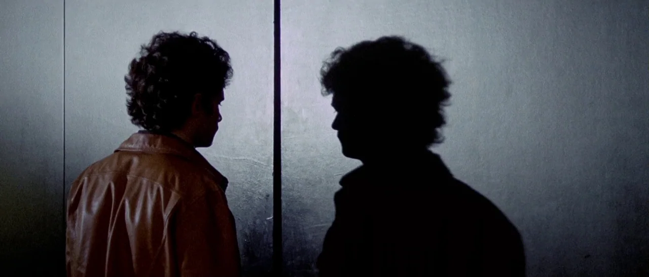 The Bird With The Crystal Plumage (1970) movie still: medium — A stark, narrow metallic space features two men standing in close proximity. The…