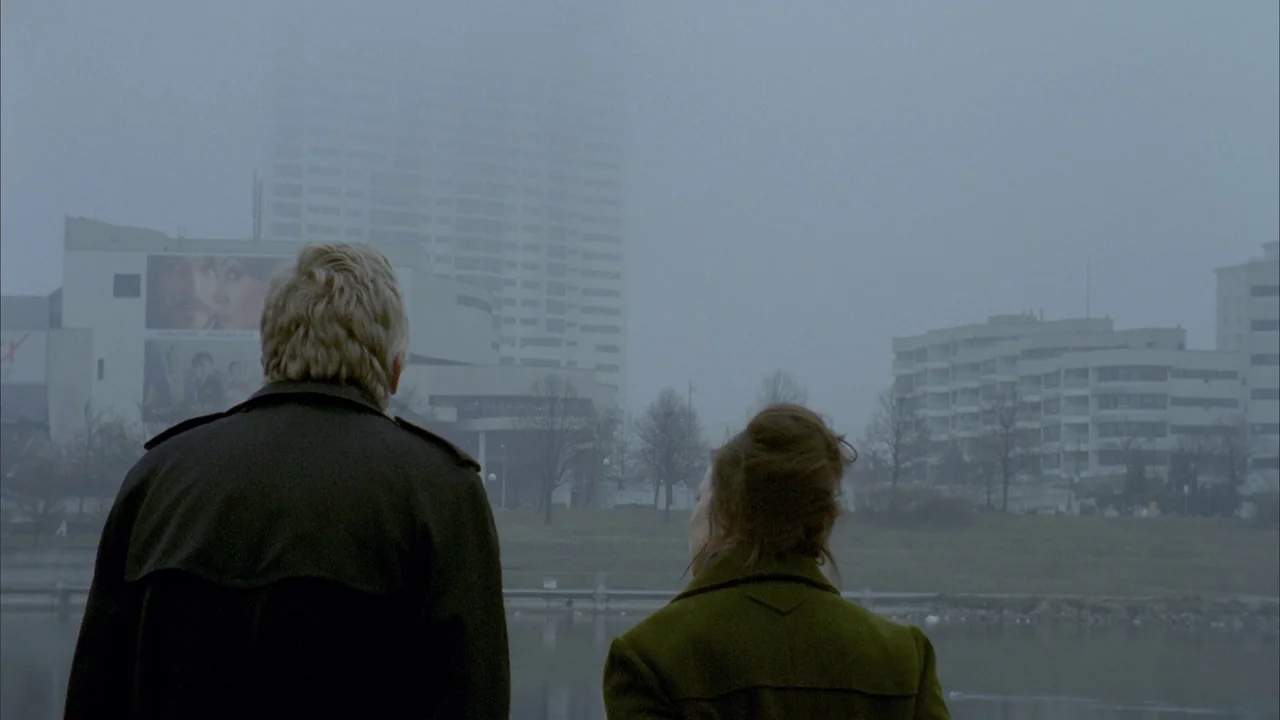 Museum Hours (2012) movie still: medium — A foggy urban riverfront with modern mid-rise buildings visible across the water. A man and a woman stand…