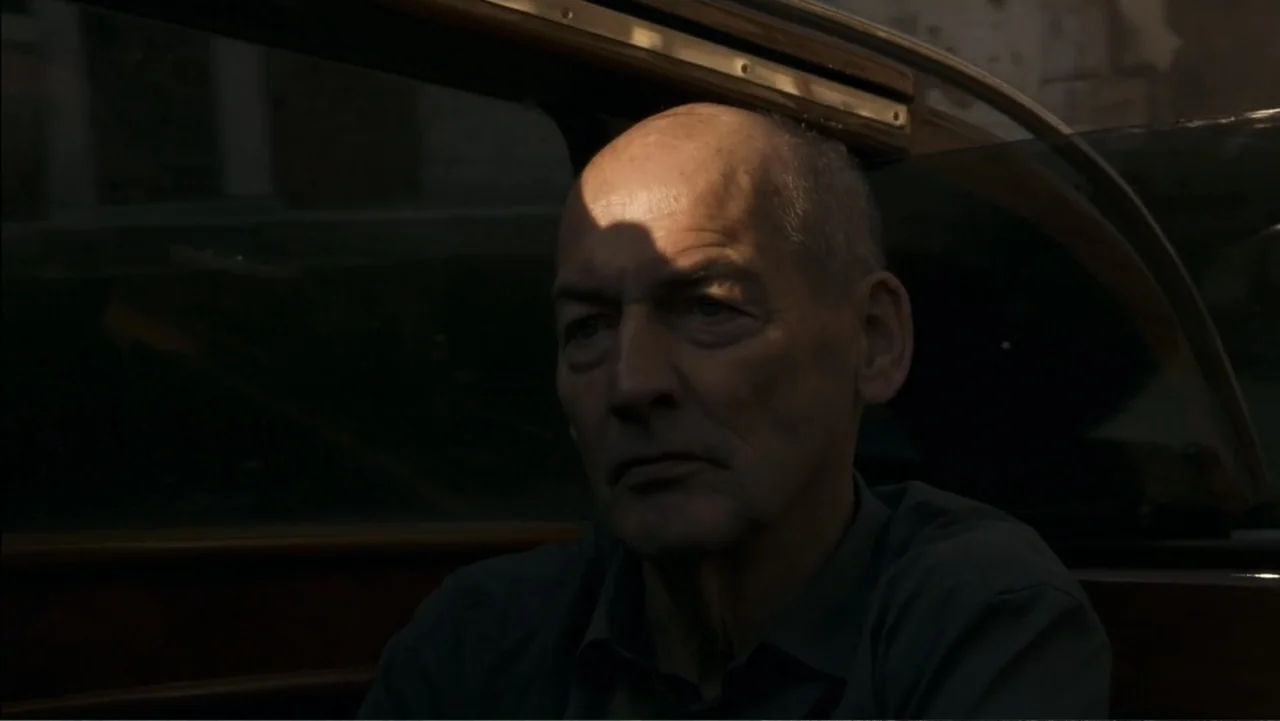 REM (2019) movie still: closeup — An older man sits inside the passenger seat of a car. He gazes straight ahead while daylight illuminates the side…