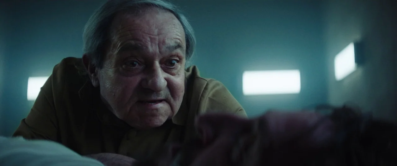 The Platform (2019) movie still: medium — A dimly lit room with rows of square wall-mounted lights glowing against dark walls. An elderly man with…