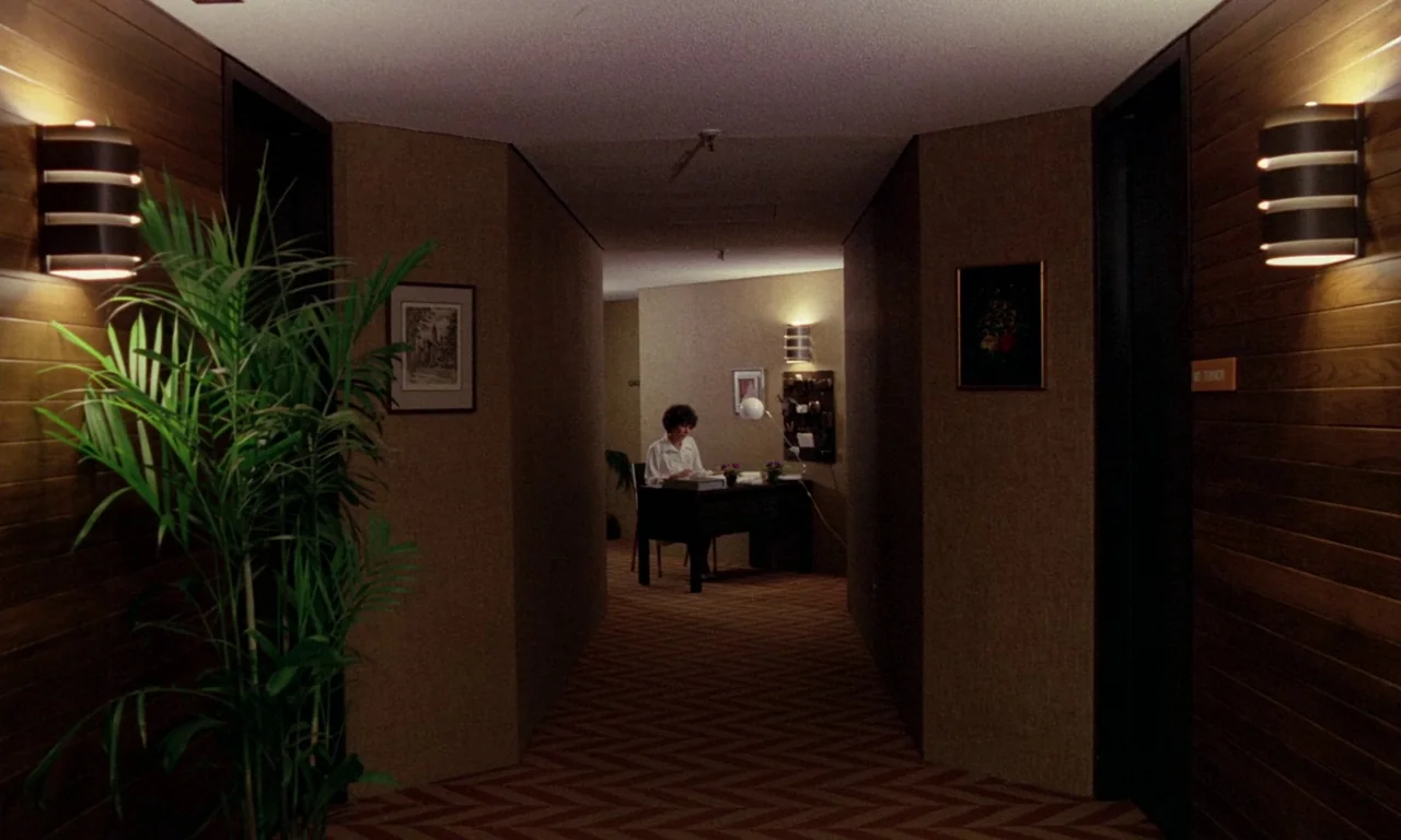 Rabid (1977) movie still: wide — A dimly lit hotel hallway with chevron-patterned carpet and dark wood-paneled walls. A woman sits at a small desk…