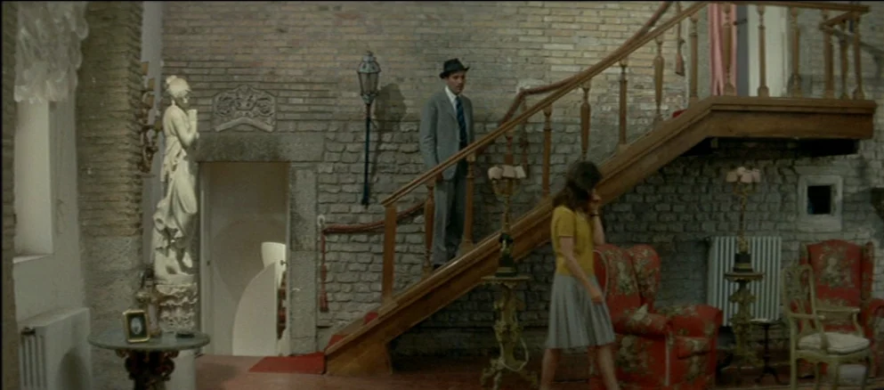 Contempt (Le Mépris) (1963) movie still: wide — A spacious room features exposed brick walls, a wooden staircase, and classical decorative statues.…