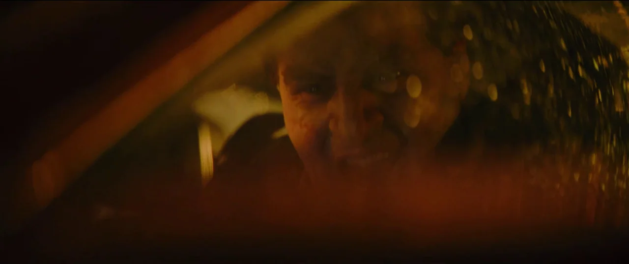 The Batman (2022) movie still: closeup — A dark vehicle interior with rain-streaked glass partially obscuring the view of the driver. A man stares…