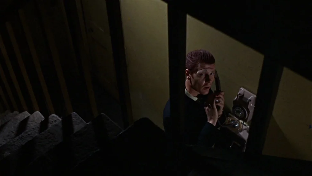 Performance (1970) movie still: medium — A dimly lit stairwell features a wall-mounted rotary telephone in a dark, confined corner. A man stands in…