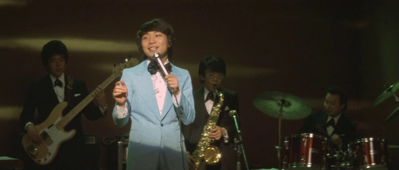 Take Me Away! (1978) movie still: medium — A musical group performs on a dimly lit stage with dark wood paneling backdrop. The lead singer stands…