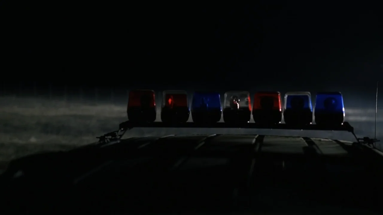 The Shepherd (2023) movie still: wide — A dark, unlit field stretches out behind the roof of a stationary police vehicle. The roof-mounted light bar…