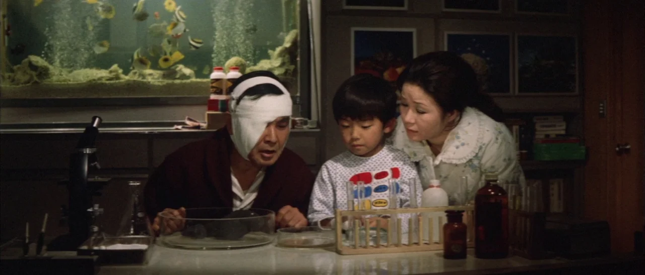 Godzilla Vs Hedorah (1971) movie still: medium — A dimly lit room with scientific equipment and a large aquarium in the back. A man with a bandaged…