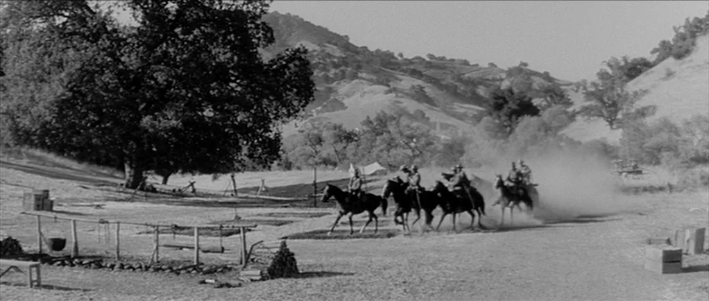 Love Me Tender movie still: wide — An open grassy field serves as a military encampment with wooden fences and equipment. Several soldiers in…