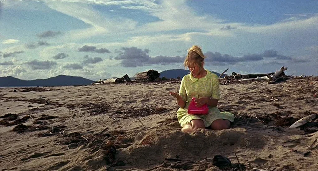 Age of Consent (1969) movie still: fullbody — A wide, debris-strewn beach meets a blue sky filled with wispy clouds and distant mountains. A young…