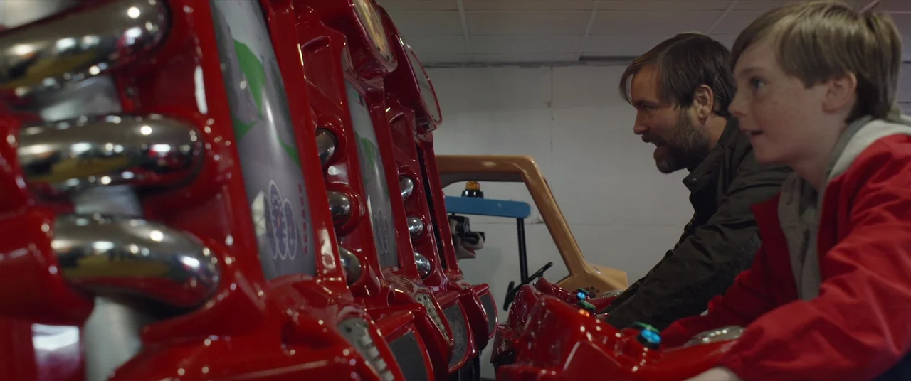 Bull (2021) movie still: medium — An indoor arcade space with multiple bright red racing cabinets lined up in a row. A man and a young boy sit at…