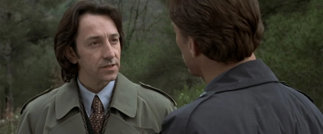 Maximum Risk (1996) movie still: medium — A wooded area with soft-focus green trees in the background during daylight hours. Two men in coats stand…