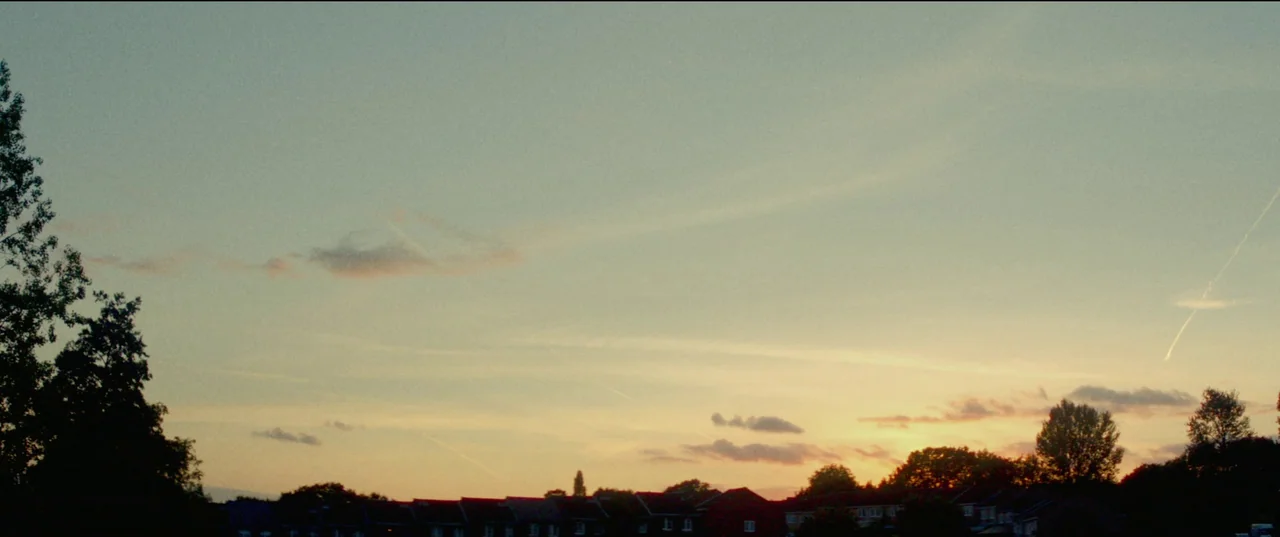 Scrapper (2023) movie still: establishing — A view of residential rooftops sits below a expansive dusk sky with thin clouds. Scattered trees frame…