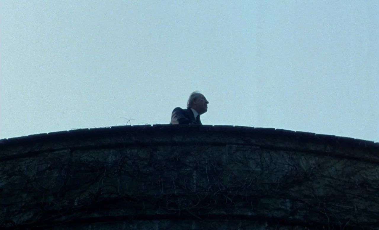 The Beast (1975) movie still: wide — An stone bridge railing covered in dried vines stands against a pale blue sky. An elderly man in a suit leans…