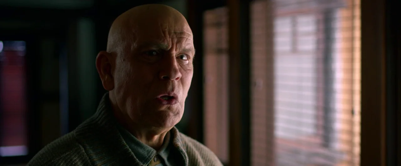 Bird Box (2018) movie still: closeup — An elderly man stands in a dark room near a window with closed blinds. He faces the camera with a surprised…