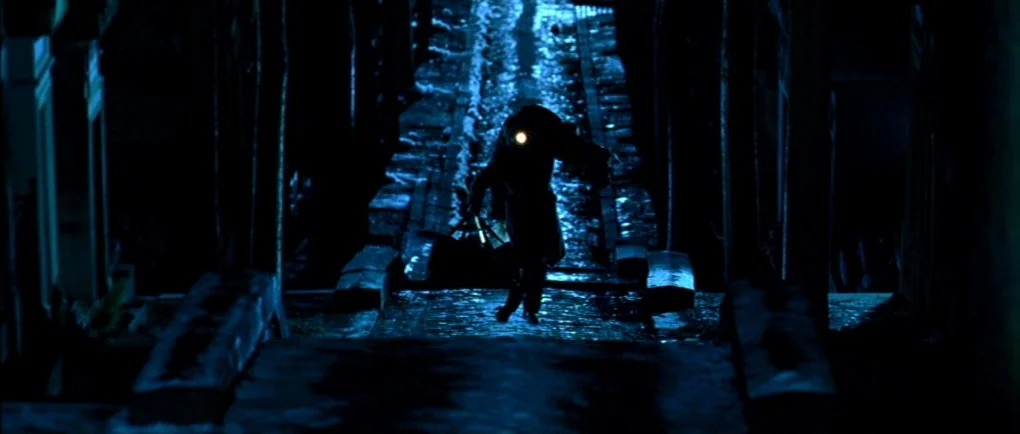 Mortal Transfer (2001) movie still: wide — A dark, narrow stone pathway at night is flanked by deep, unlit vertical structures. A single silhouetted…