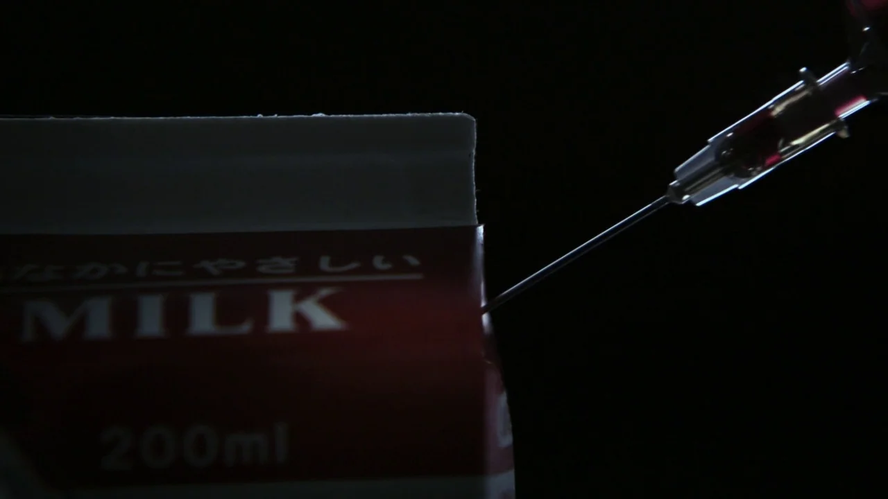 Confessions (2010) movie still: closeup — A close-up shot focuses on a small cardboard milk carton against an underexposed background. A metal…