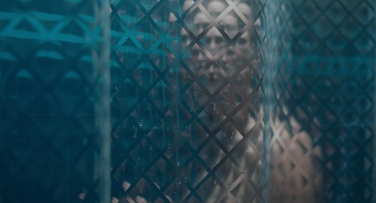 La Abuela (2021) movie still: closeup — A person stands behind a textured diamond-patterned glass shower door in a dim room. The light filters…