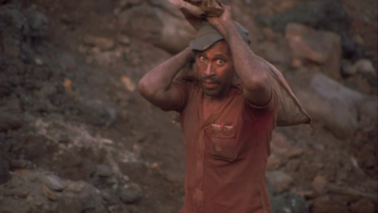 Powaqqatsi (1988) movie still: medium — A man works in a dirt-filled area surrounded by mounds of dark earth. He carries a heavy sack on his head…