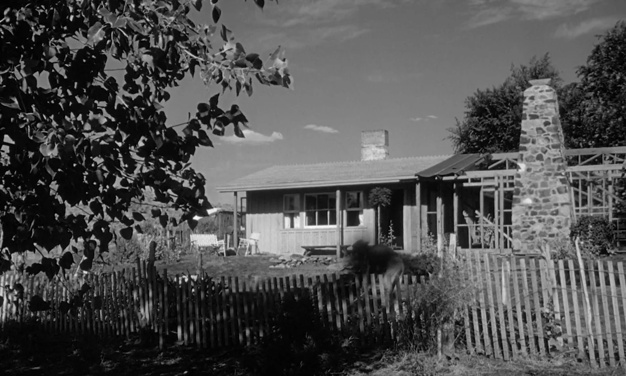 The Misfits (1961) movie still: wide — A rural farmhouse sits behind a weathered wooden picket fence in a grassy yard. A stone chimney stands on the…