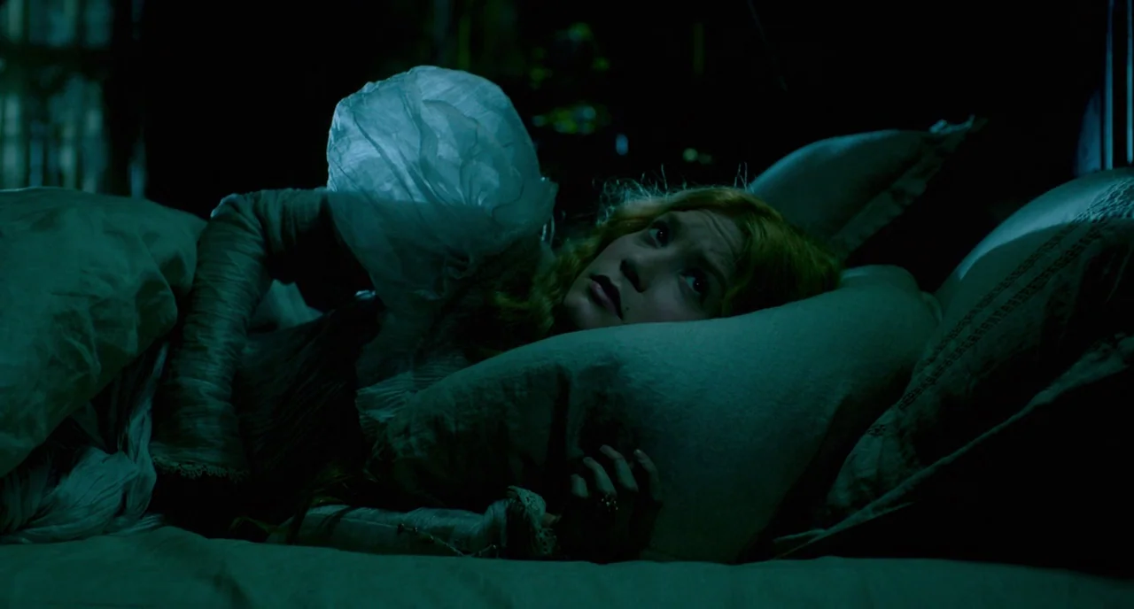 Crimson Peak (2015) movie still: closeup — A dark bedroom setting with a person lying in a bed with white linens. The subject rests her head on a…