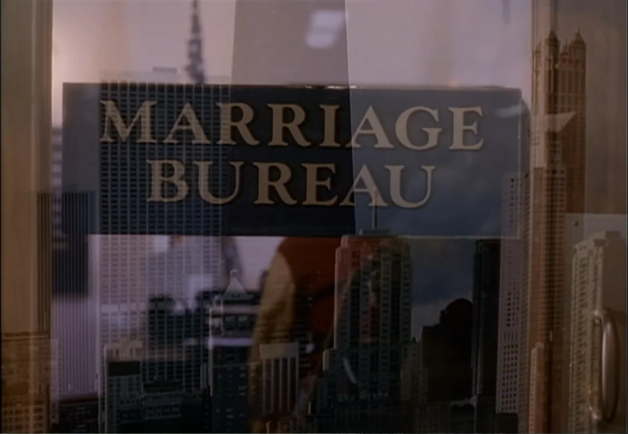 Chance of a Lifetime (1998) movie still: medium — A glass door features a Marriage Bureau sign with a city skyline reflection overlay. A person…