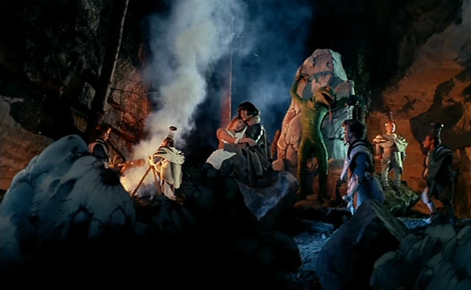 Life Is A Bed of Roses (1983) movie still: wide — A dark, rocky cave interior features a small campfire producing thick plumes of smoke. A group of…