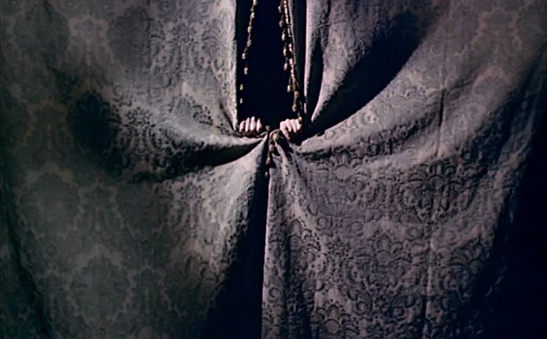 Mill of the Stone Women (1960) movie still: closeup — A dark space is partially concealed by a heavy fabric curtain with a damask pattern. A pair of…