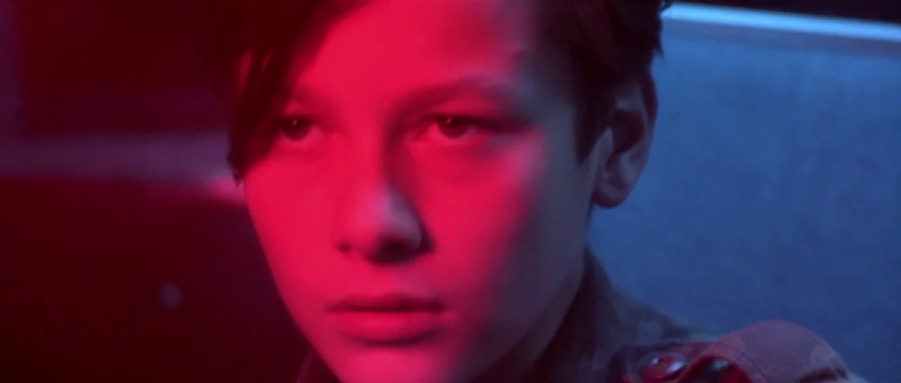 Terminator 2: Judgement Day (1991) movie still: closeup — The face of a young male teenager is illuminated by sharp red and blue lights. He looks…