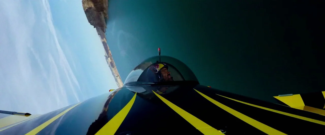 6 Underground (2019) movie still: medium — An acrobatic airplane flies inverted over a body of water with high cliffs nearby. The pilot sits in the…