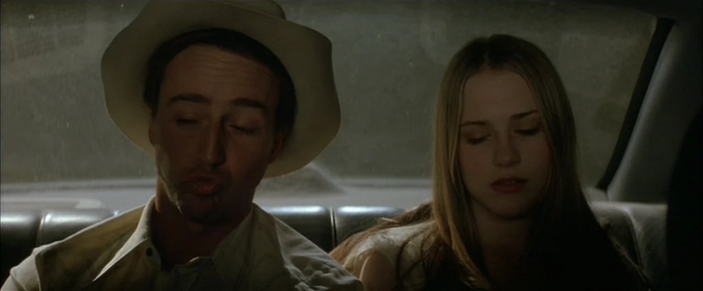 Down In The Valley (2005) movie still: medium — Two individuals sit in the dark backseat of a moving car at night. The man in a hat sits beside a…