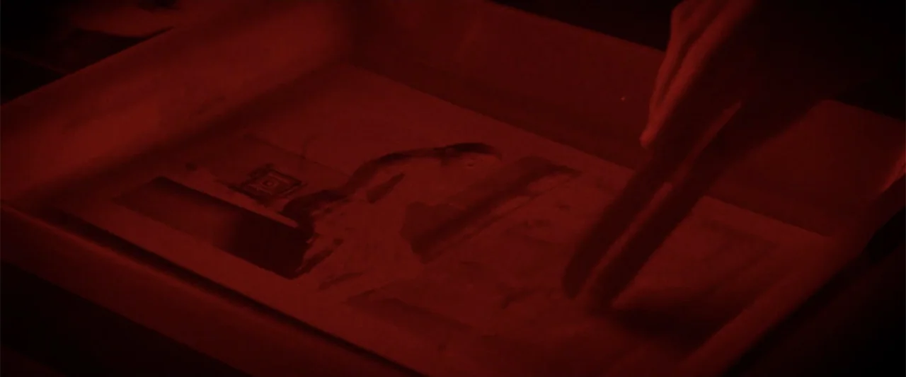 Anna (2013) movie still: closeup — A darkroom tray contains a photographic print partially submerged in developing chemicals. A human hand moves the…